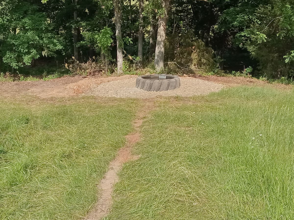 A tire tank on a gravel pad at the edge of a pasture, with the healed line of the waterline run visible crossing the grass toward it