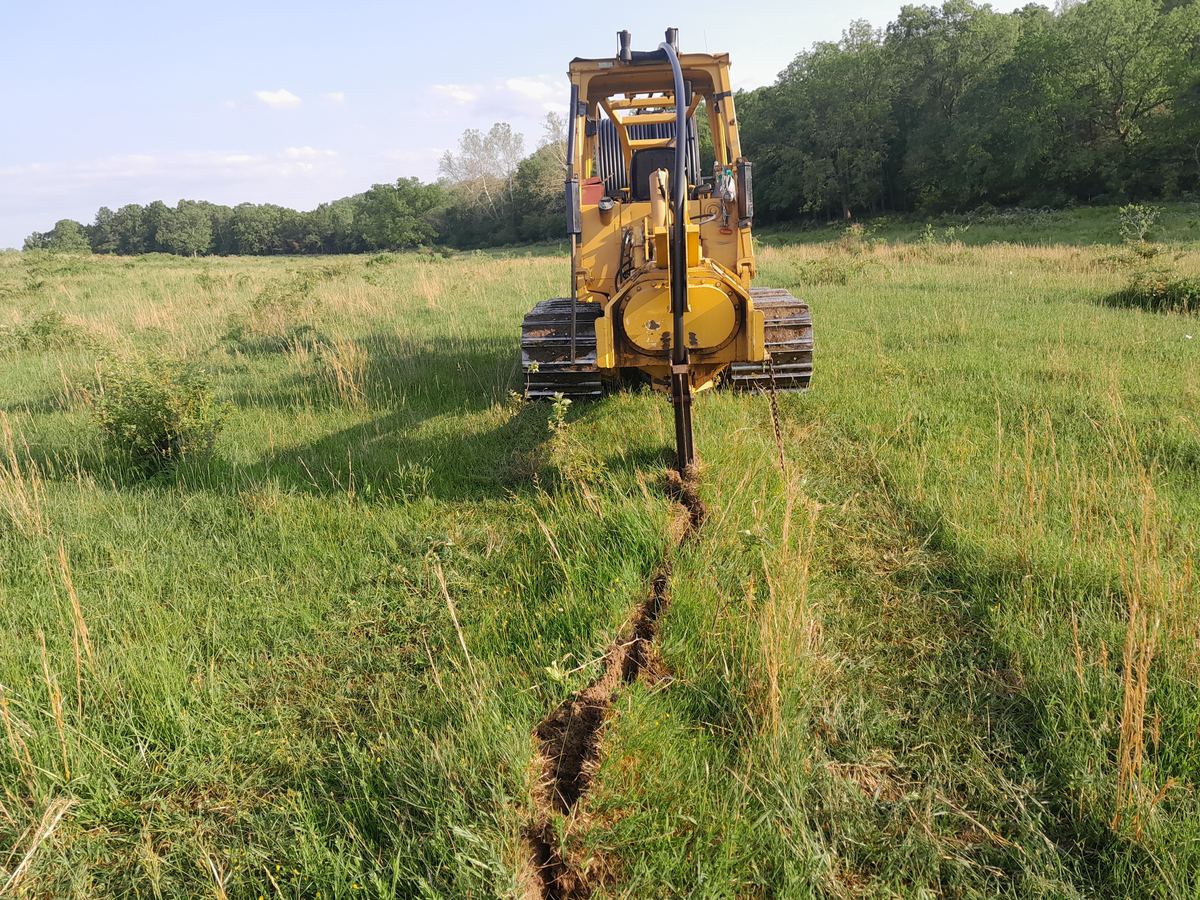 A vibratory plow pulling waterline through standing pasture, leaving a narrow slit in the sod behind it instead of an open trench