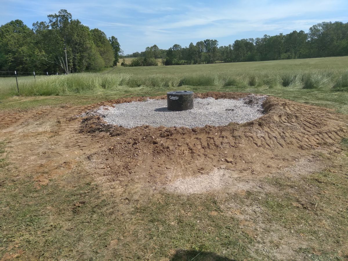 A finished watering tank standing on a graded gravel pad in open pasture, with the fence line and tree line of the farm behind it